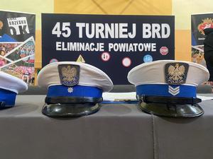 czapki policyjne z bialym otokiem położone na stoliku, w tle baner z napisem 45 turniej bezpieczeństwa w ruchu drogowym