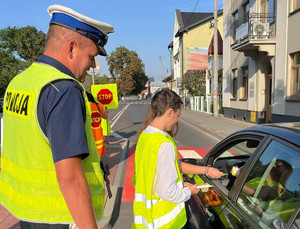 uczniowie w towarzystwie policjanta wręczają ulotkę zwalniam przed szkołą kierowcy