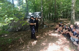 policjantki z grupą młodzieży podczas prelekcji przy obozowisku w lesie, w tle namiot