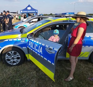 dziecko siedzące w radiowozie, obok radiowozu kobieta, w tle policjanci