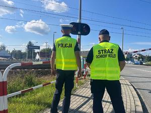policjant i przedstawiciel straży ochrony kolei stojący tyłem przy przejeździe kolejowym