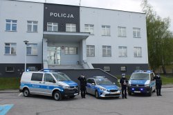 Na zdjęciu znajdują się trzy radiowozy Policji, po prawej i lewej stronie widoczne są furgony policyjne po środku stoi radiowóz osobowy. Wszystkie pojazdy posiadają włączone sygnały błyskowe. Obok radiowozów stoją umundurowani funkcjonariusze. Policjant stojący na środku trzyma w ręku znicz. W tle widać budynek Komendy Powiatowej Policji w Brzesku.