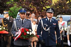 Komendant Powiatowy Policji w Brzesku wraz z policjantem niosącym biało-czerwoną wiązankę kwiatów, w tle uczestnicy uroczystości