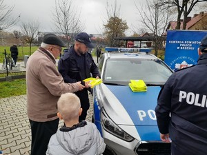 policjant wręczający kamizelkę odblaskową mężczyźnie z dzieckiem, w tle radiowóz