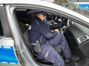 policjant siedzący  na miejscu pasażera pokazujący wyposażenie radiowozu dziecku siedzącemu na fotelu kierowcy