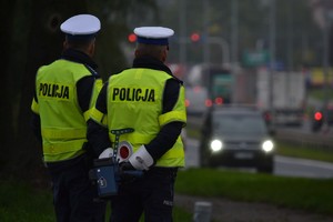 Dwóch policjantów ruchu drogowego stoi przy drodze, nadzoruje ruch. Jeden z policjantów tryma za sobą ręczny miernik prędkości. W tle widać nadjeżdżające auto