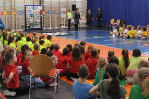 dzieci siedzące na podłodze auli w trakcie rozpoczęcia turnieju, w tle dyrektor szkoły i organizatorzy