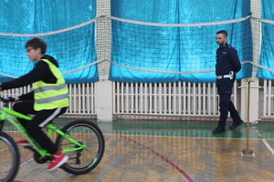 uczeń pokonujący tor przeszkód na rowerze, w tle policjant sprawdzający poprawność wykonania toru