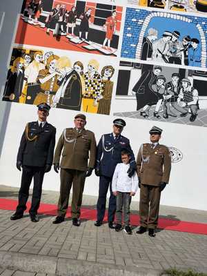przedstawiciele służb mundurowych policji, straży pożarnej, wojska z chłopcem, w tle odsłonięty mural