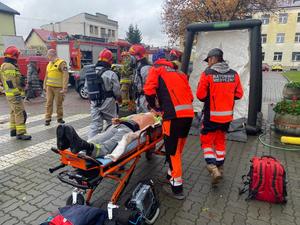 ratownicy medyczni udzielający pomocy poszkodowanemu leżącemu na noszach, w tle strażacy
